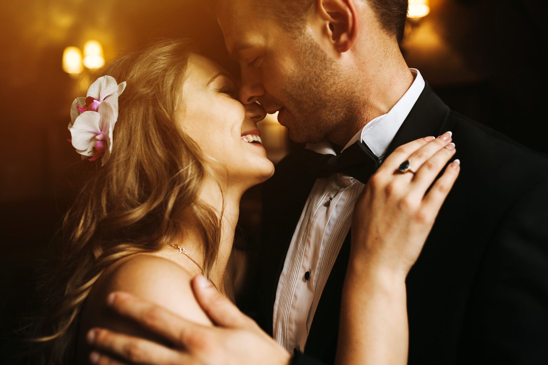 Una pareja vestida de etiqueta se abraza y sonríe con complicidad en un ambiente cálido y tenuemente iluminado.