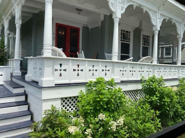 White Victorian house with porch; railing with crosses, red door, green bushes.