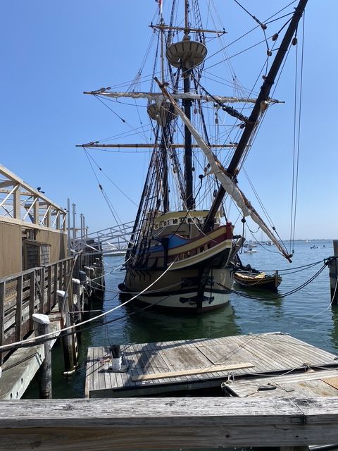 Tall ship docked at pier on a sunny day. Blue water, clear sky. Wooden dock in foreground.