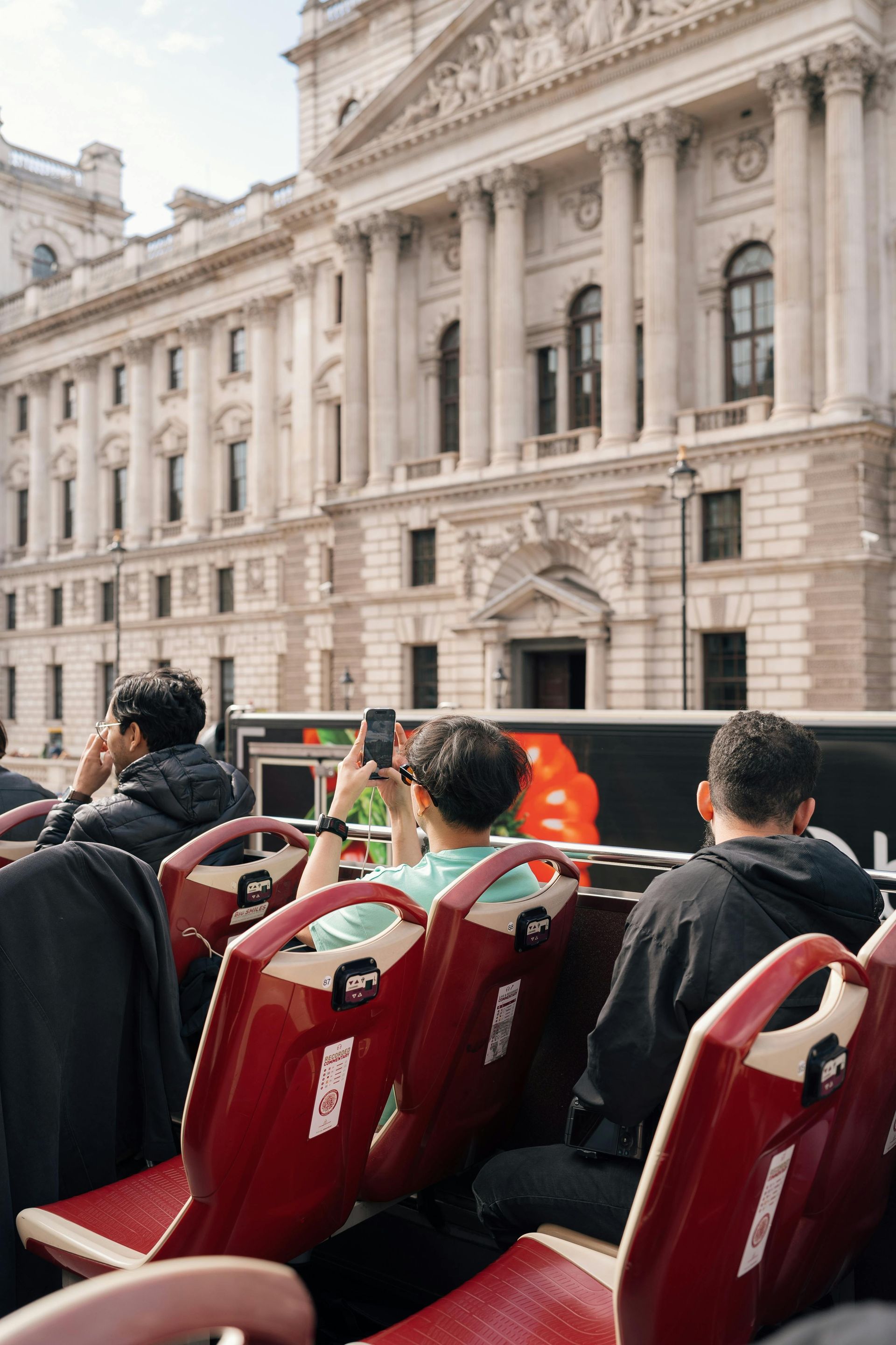 People on a red open-top bus, taking photos in front of a classical building.