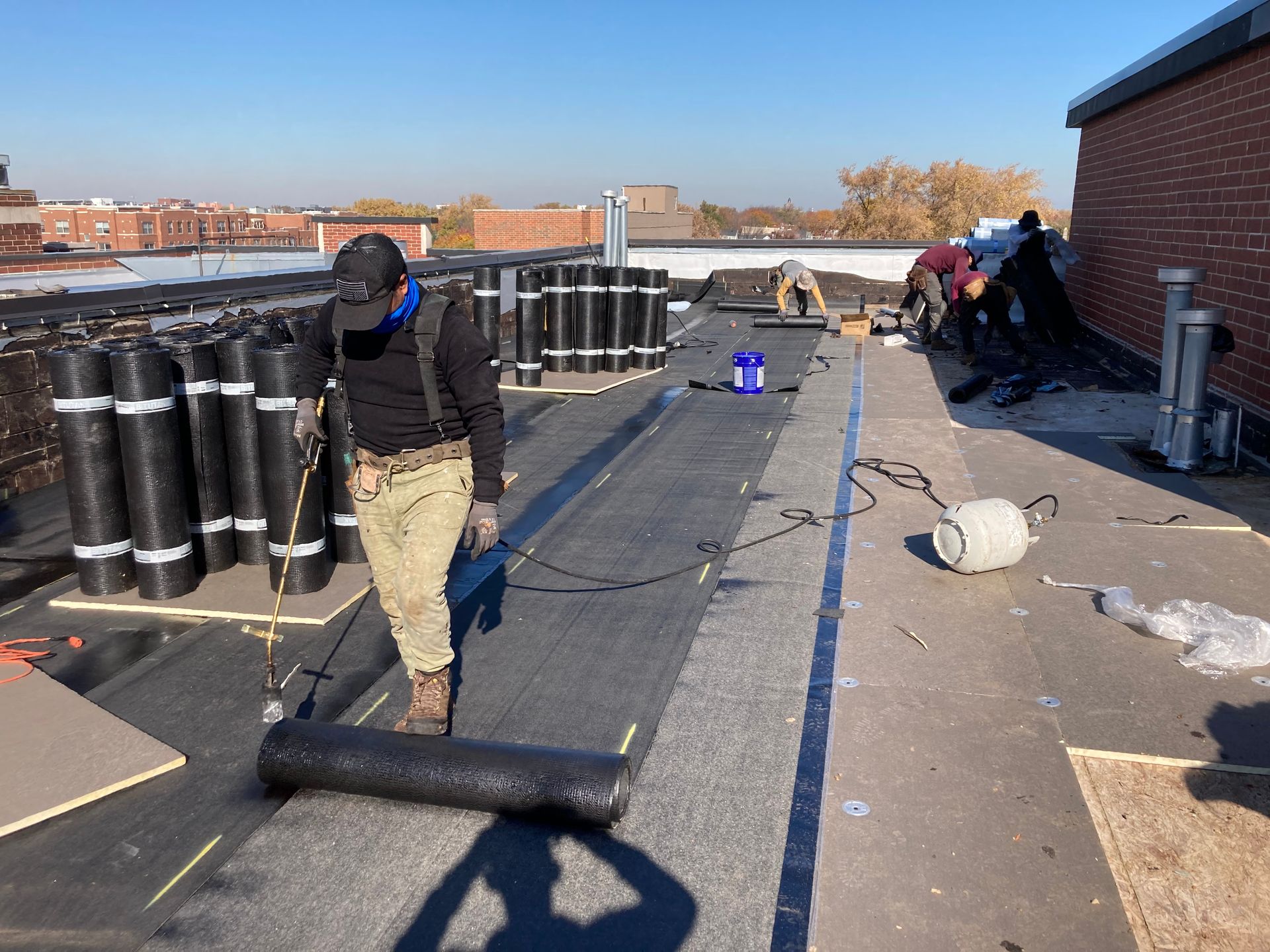 Roofers installing black roofing material on a flat rooftop. Rolls of roofing and equipment visible. Sunny day.