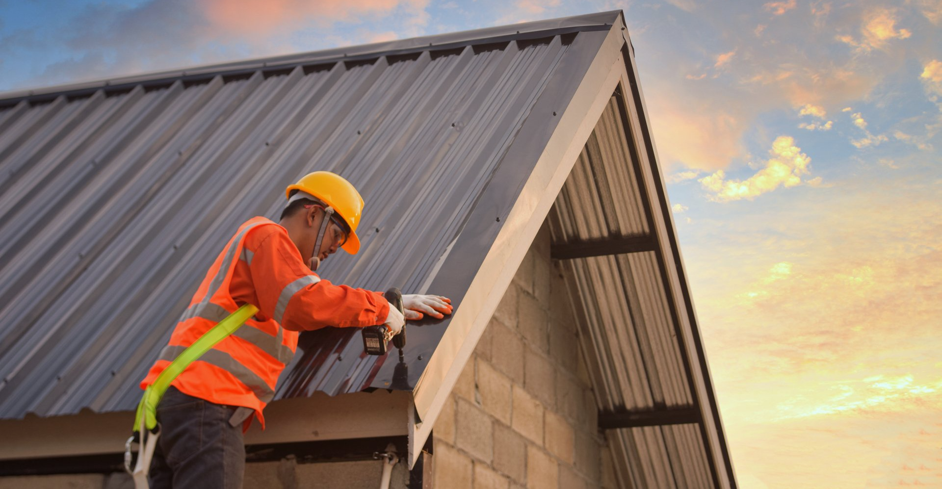 Roofer in safety gear installing metal roof panels on a building under a sunset sky.