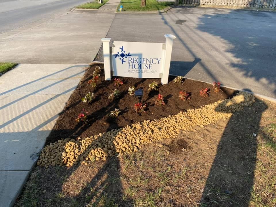 Flower bed with a small memorial sign beside a sidewalk, bordered by mulch and fresh plants.