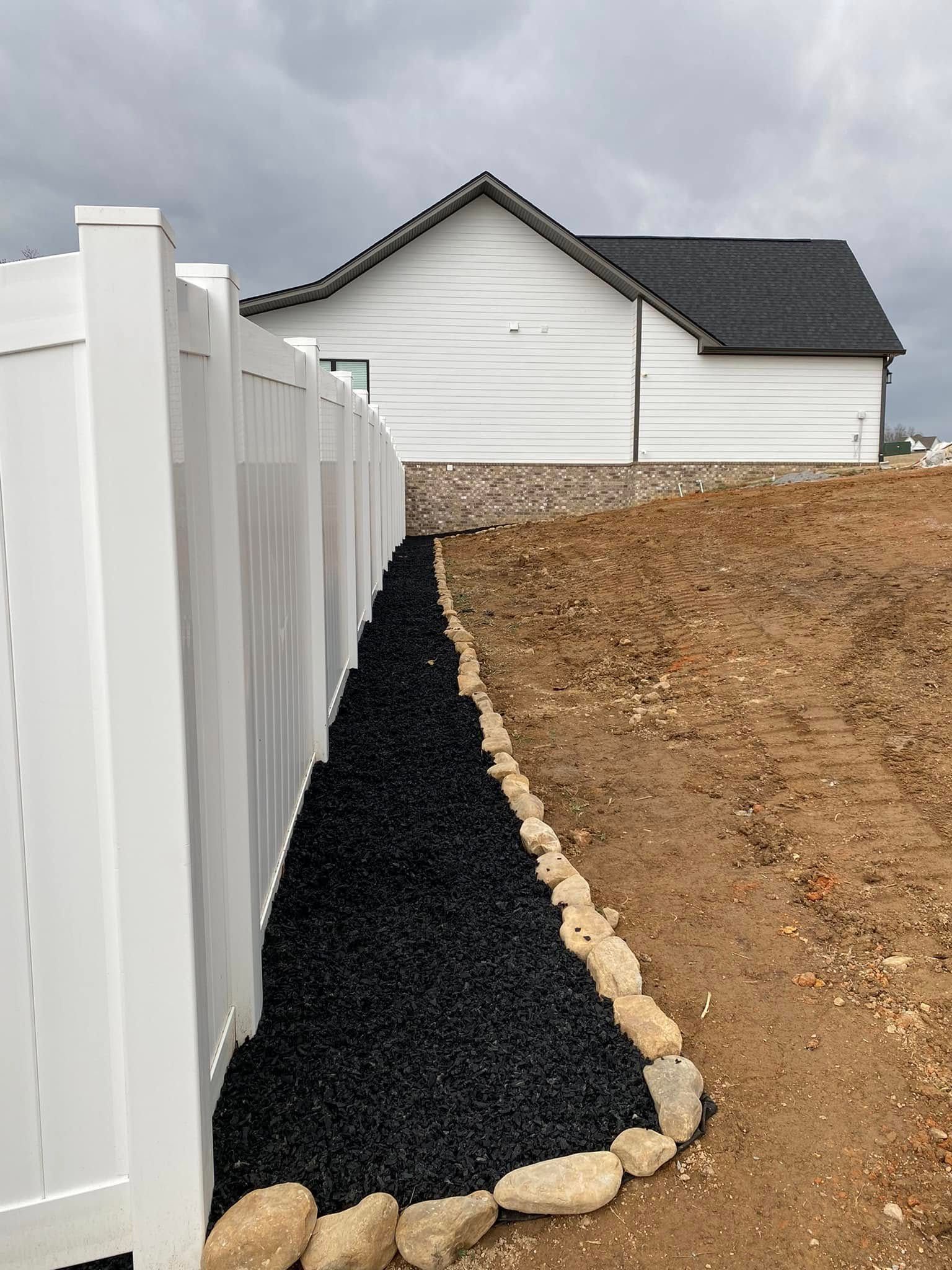 White fence beside black gravel path and tan dirt beside a white house under cloudy sky