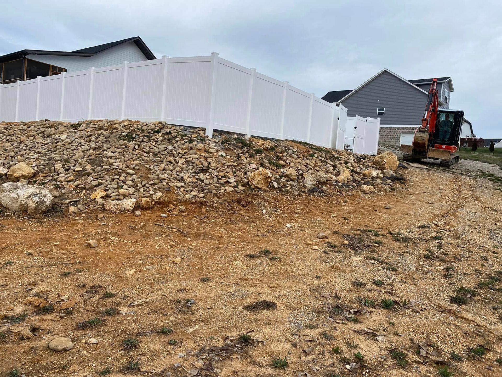 Construction site with white fence, rocky dirt slope, houses, and an excavator on the right