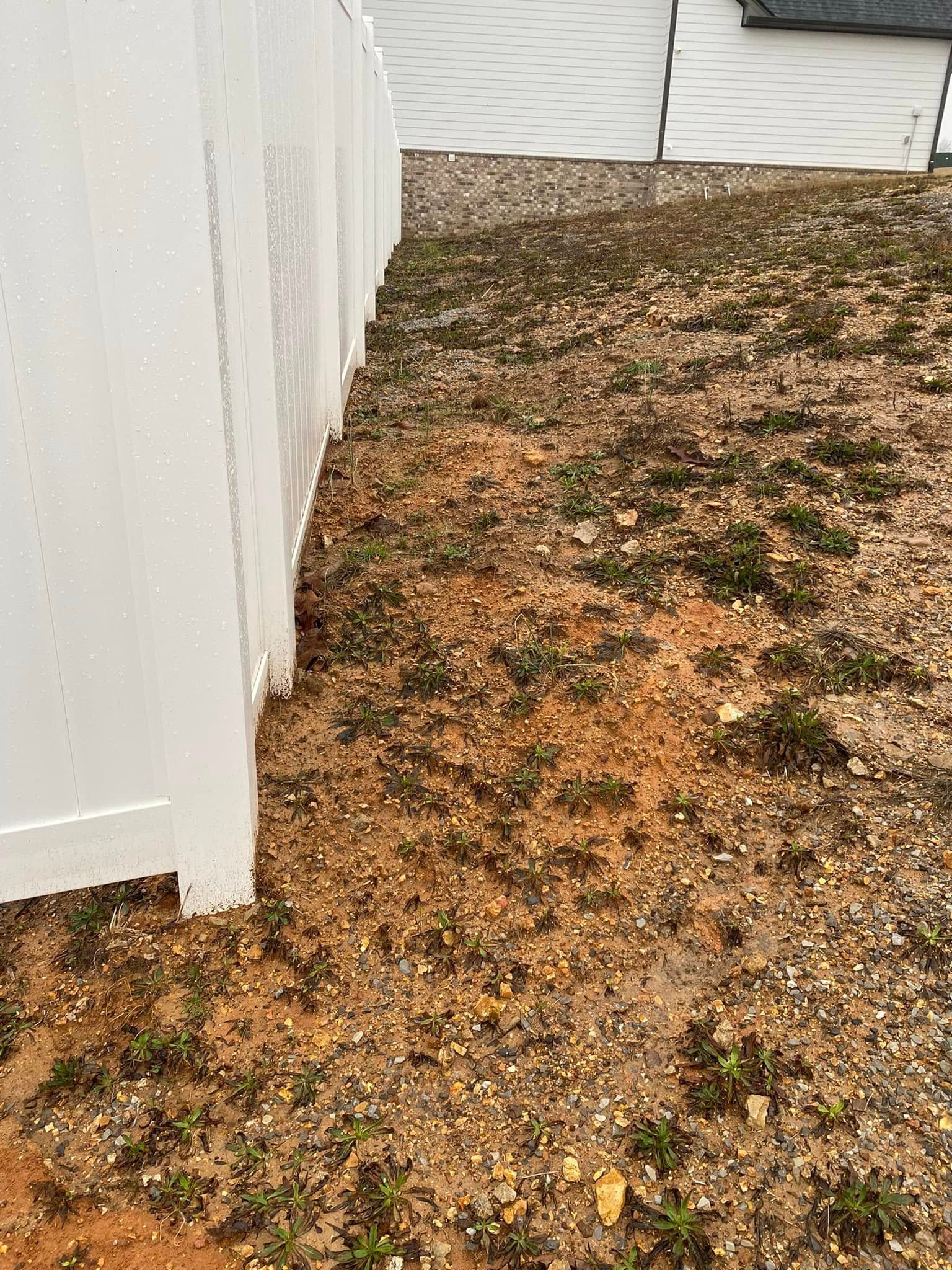 Narrow gravel strip beside a white fence and house, with patchy grass and bare soil.