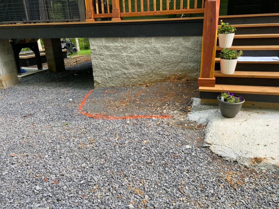 Gravel under a raised wooden deck with stone wall, orange spray marking, and potted plants by steps