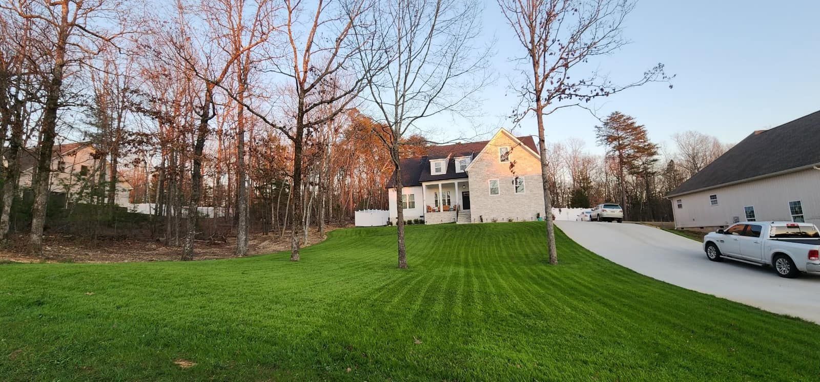 Suburban house beside a green lawn and driveway at sunset, with trees and parked cars nearby.