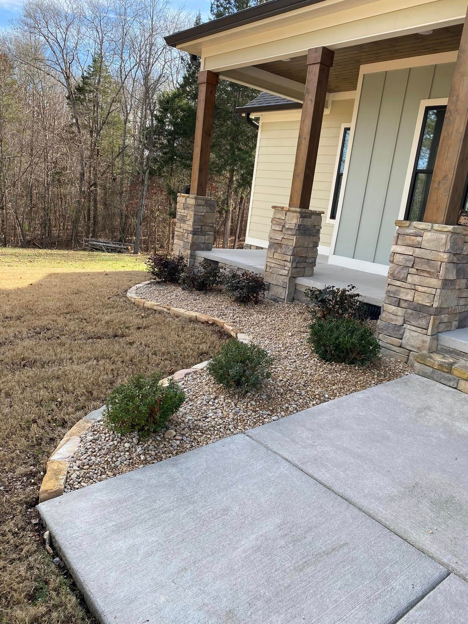 Front porch of a beige house with stone columns, concrete walkway, and landscaped mulch beds