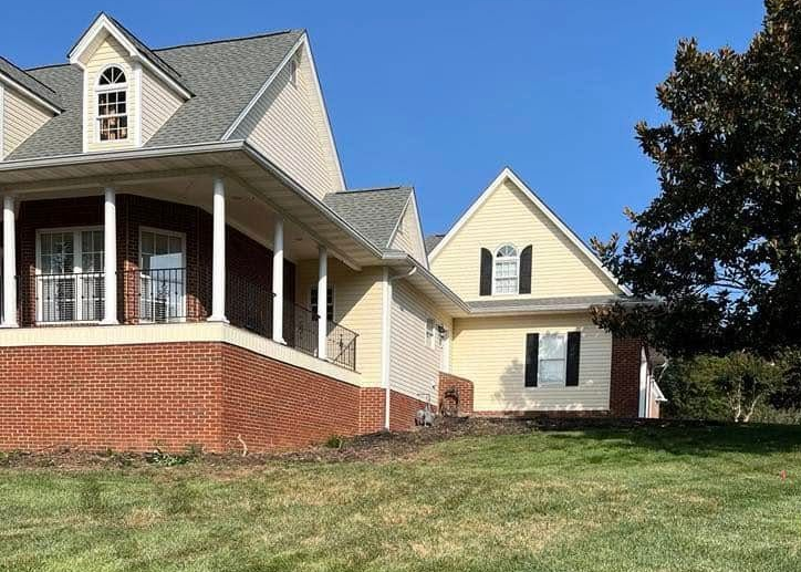 Two-story beige house with red brick porch and white columns on a grassy hill under a clear blue sky