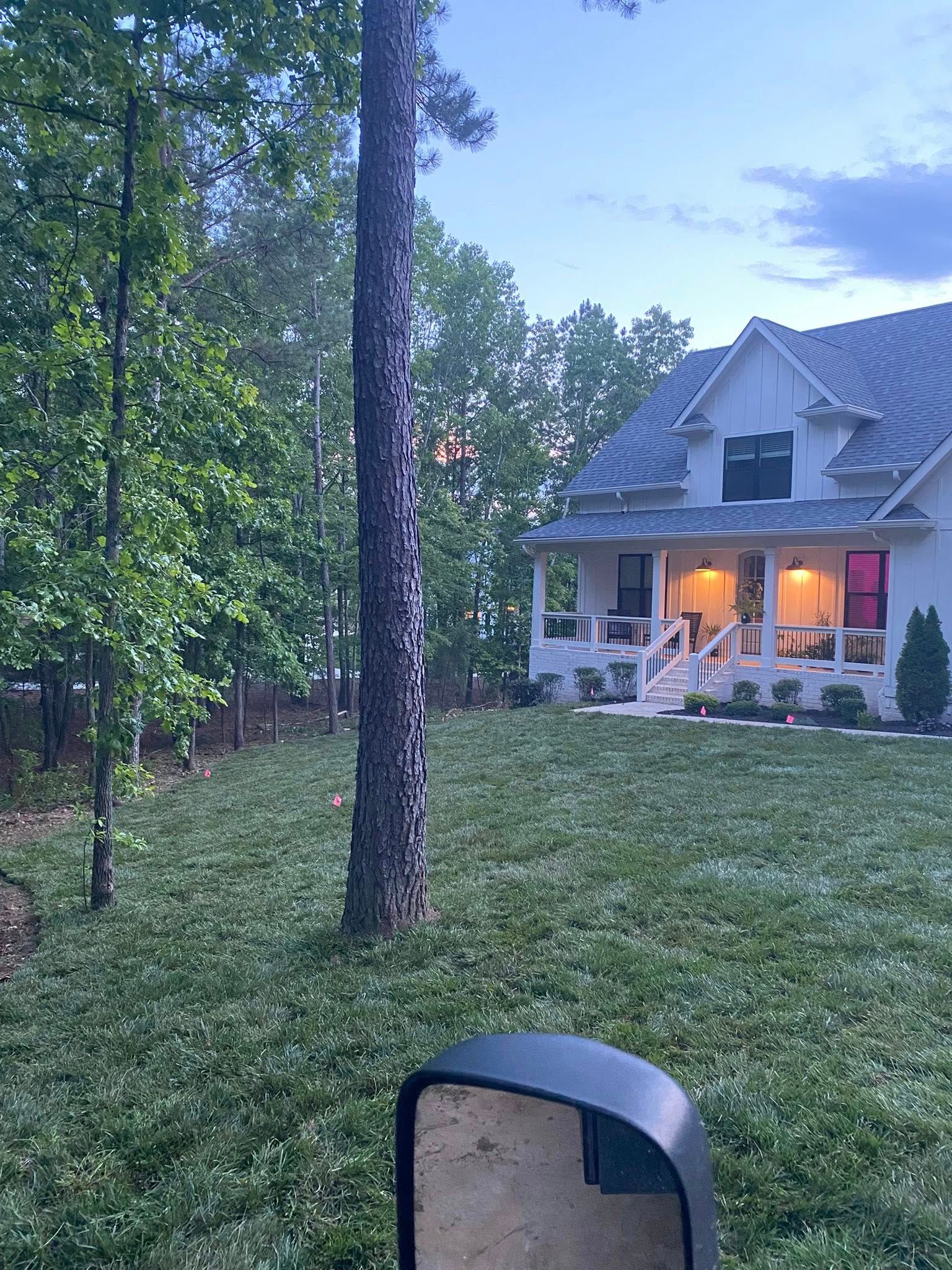 House with lit porch at dusk, surrounded by trees and a grassy yard.