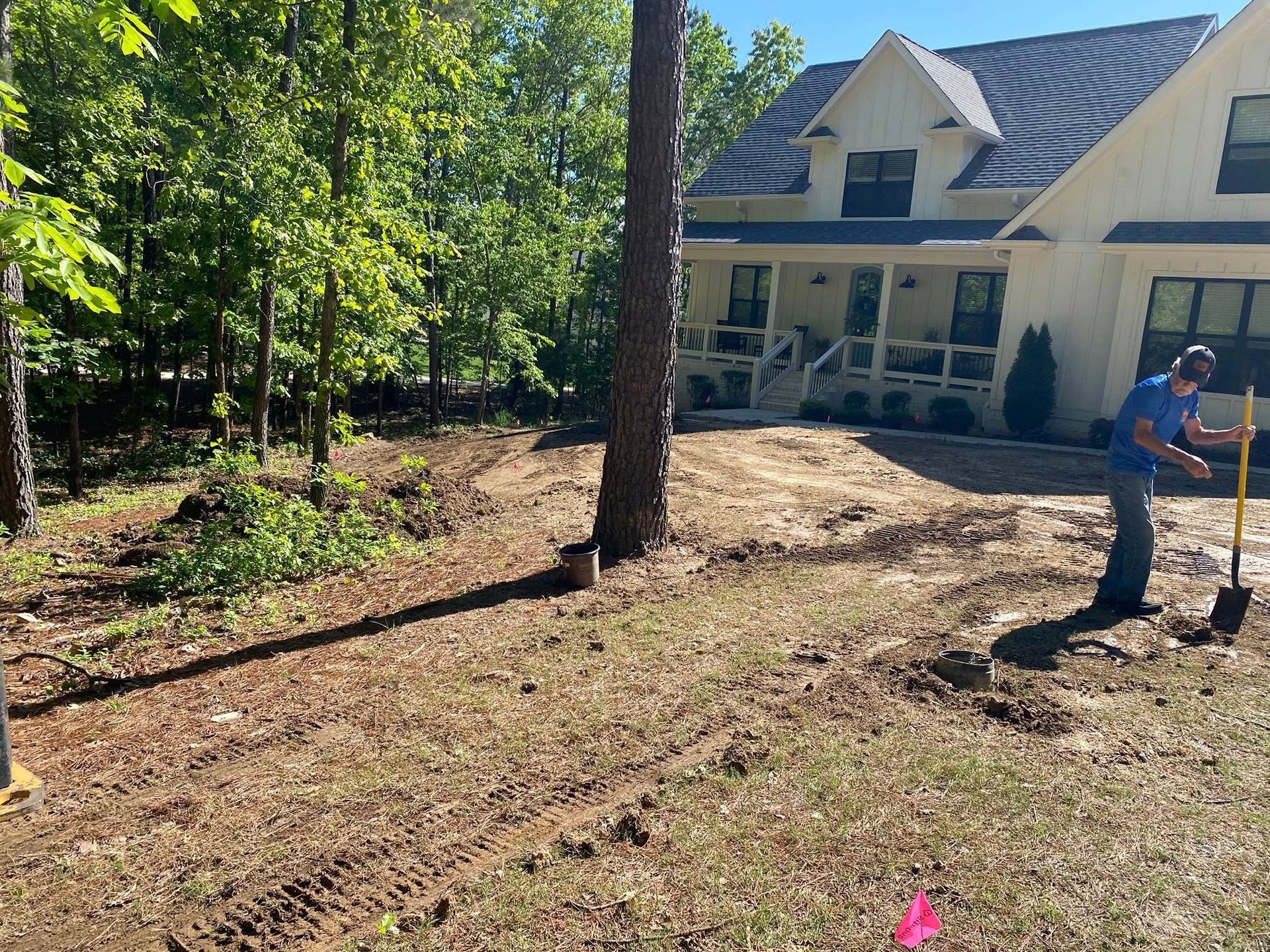 Yard work beside a house, with a person using a rake near a tree and a cleared dirt area.