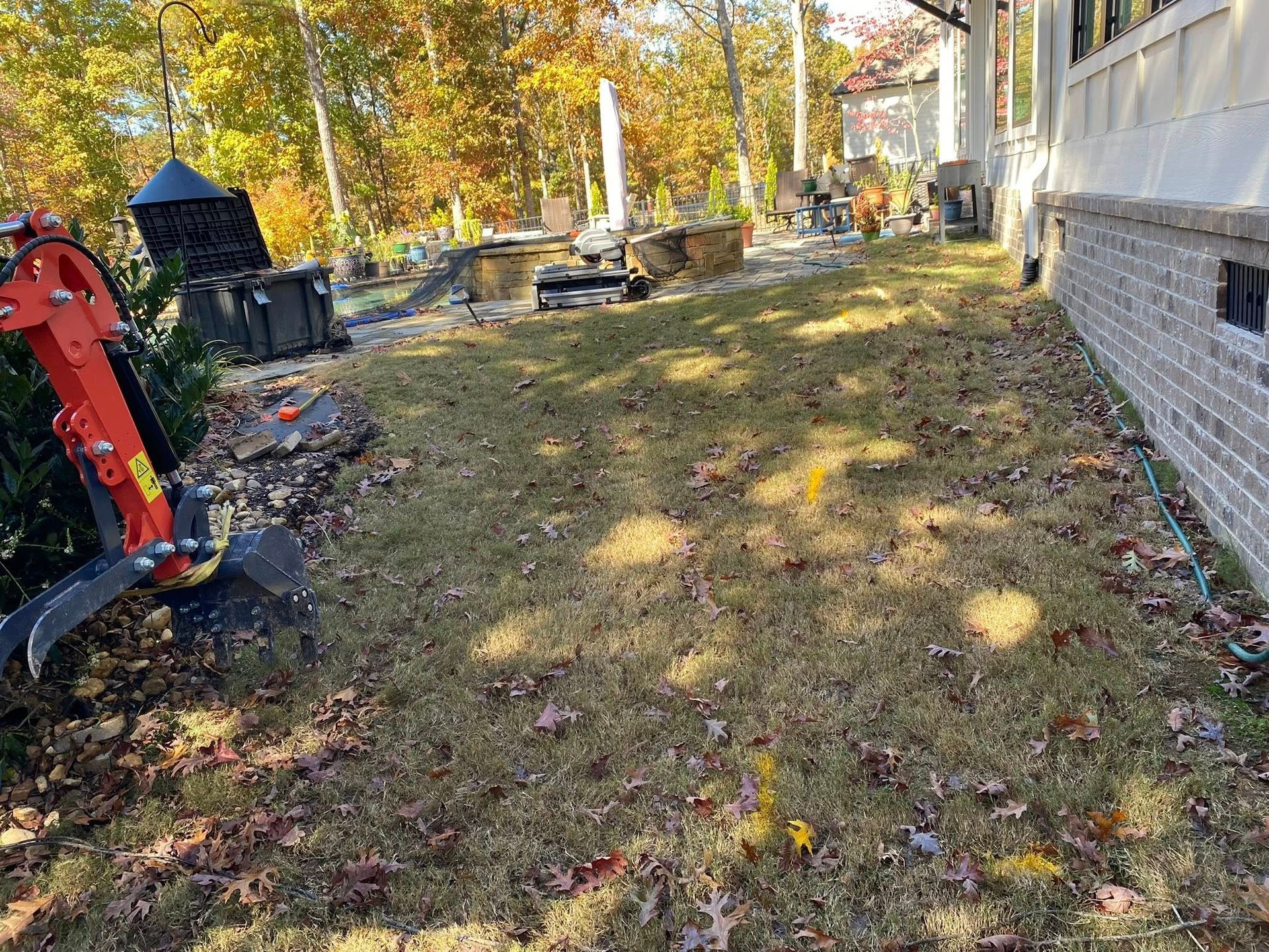 Leaf-covered yard beside a house, with a red excavator on the left and autumn trees in the background