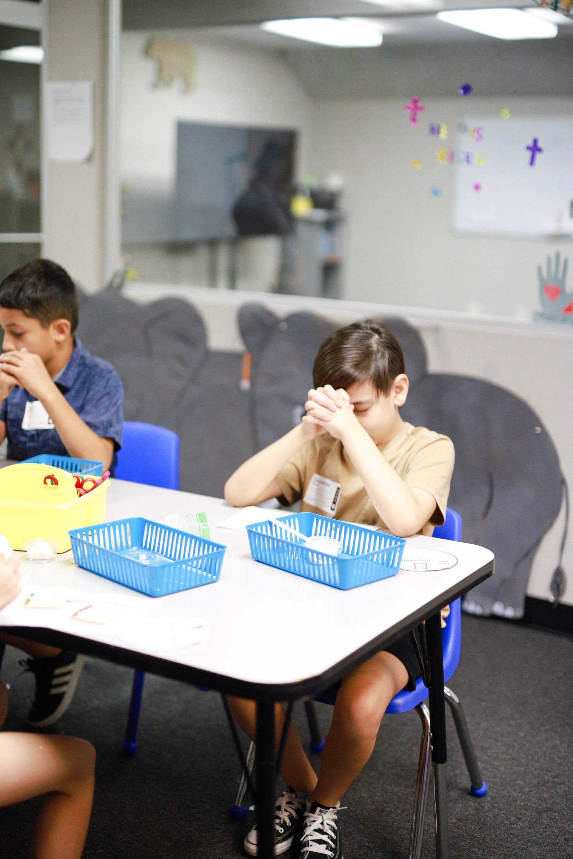 Two boys at a table, one praying with hands clasped, the other eating. Blue chairs, white table. Classroom setting.