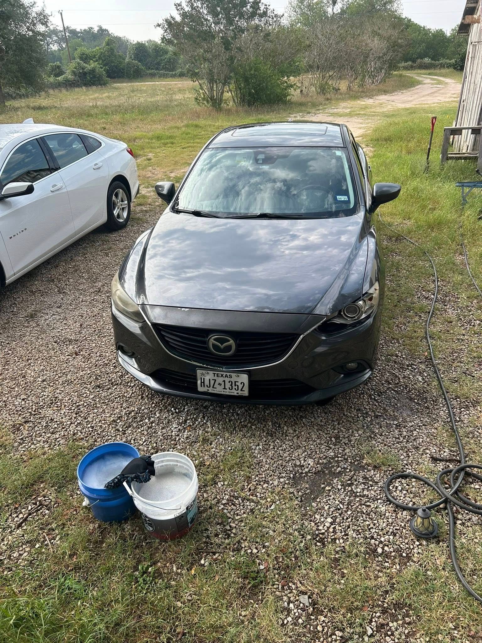 Black sedan parked on gravel beside a white car, with two buckets in the foreground and trees behind.