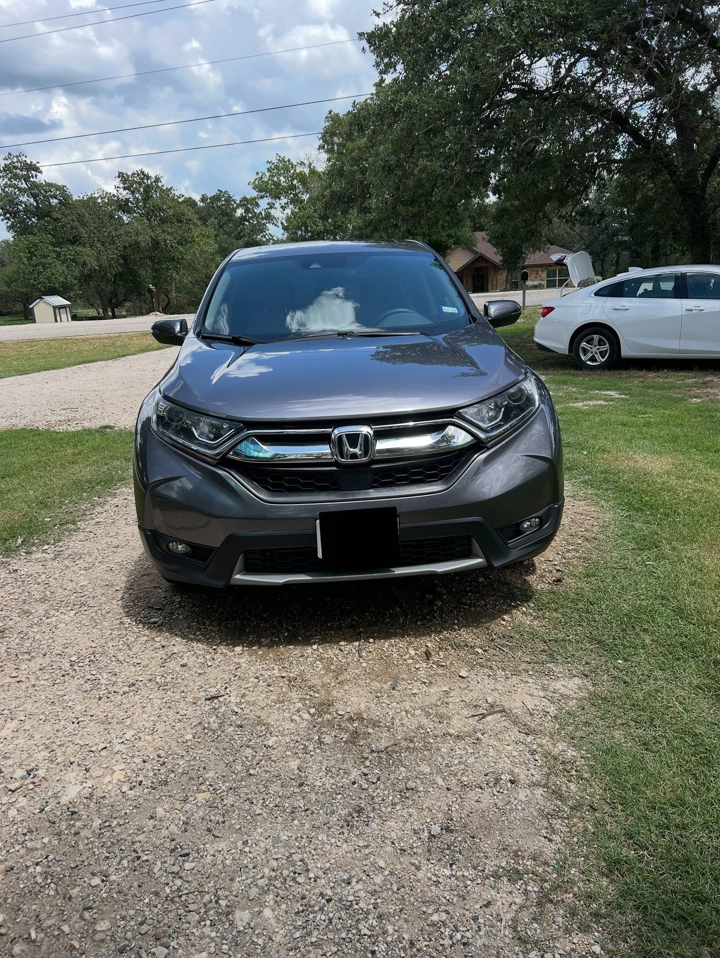 Front view of a dark gray Honda SUV parked on a gravel driveway near other cars and trees.