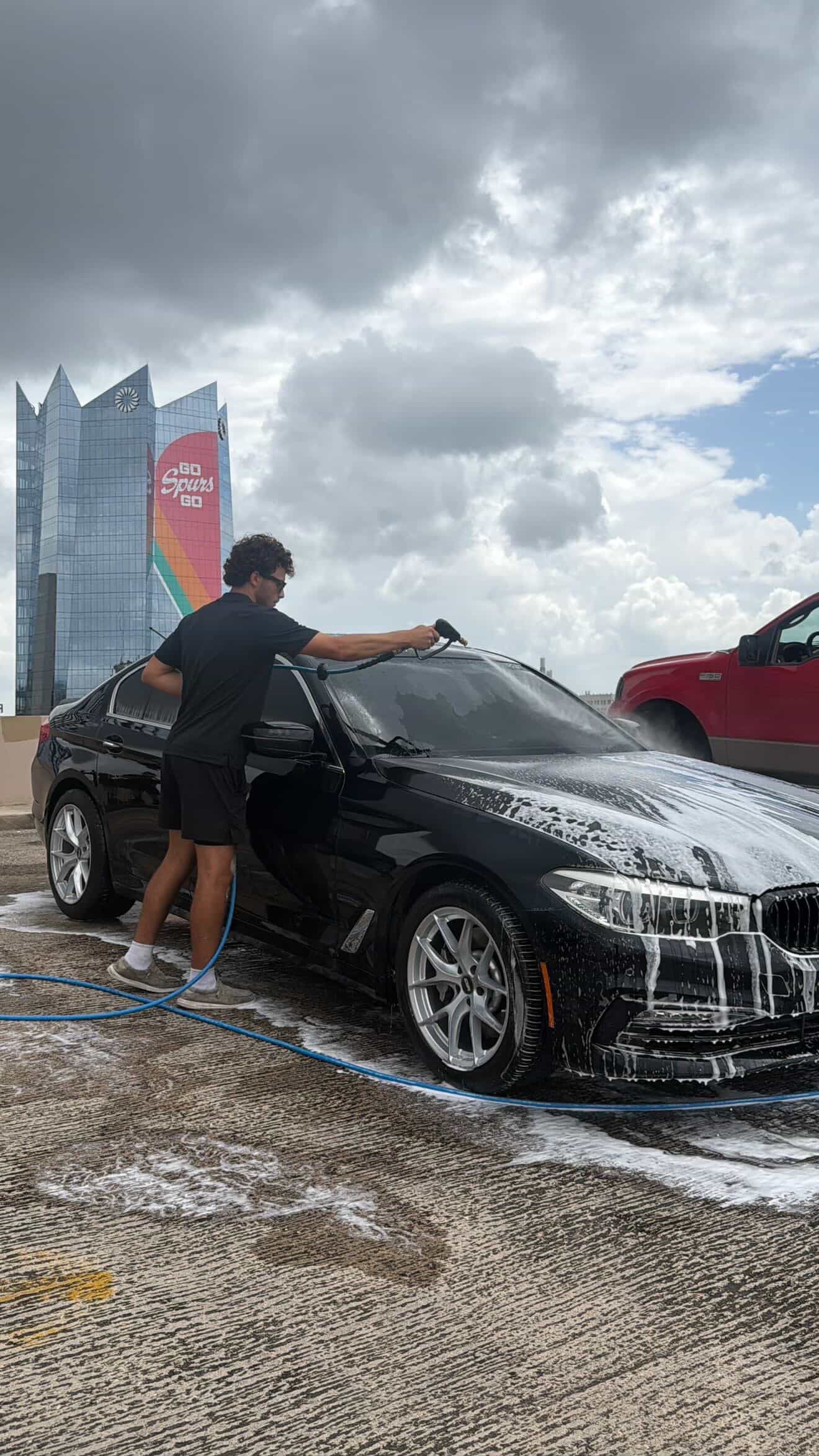 Person washing a black sports car with foam outdoors under cloudy skies