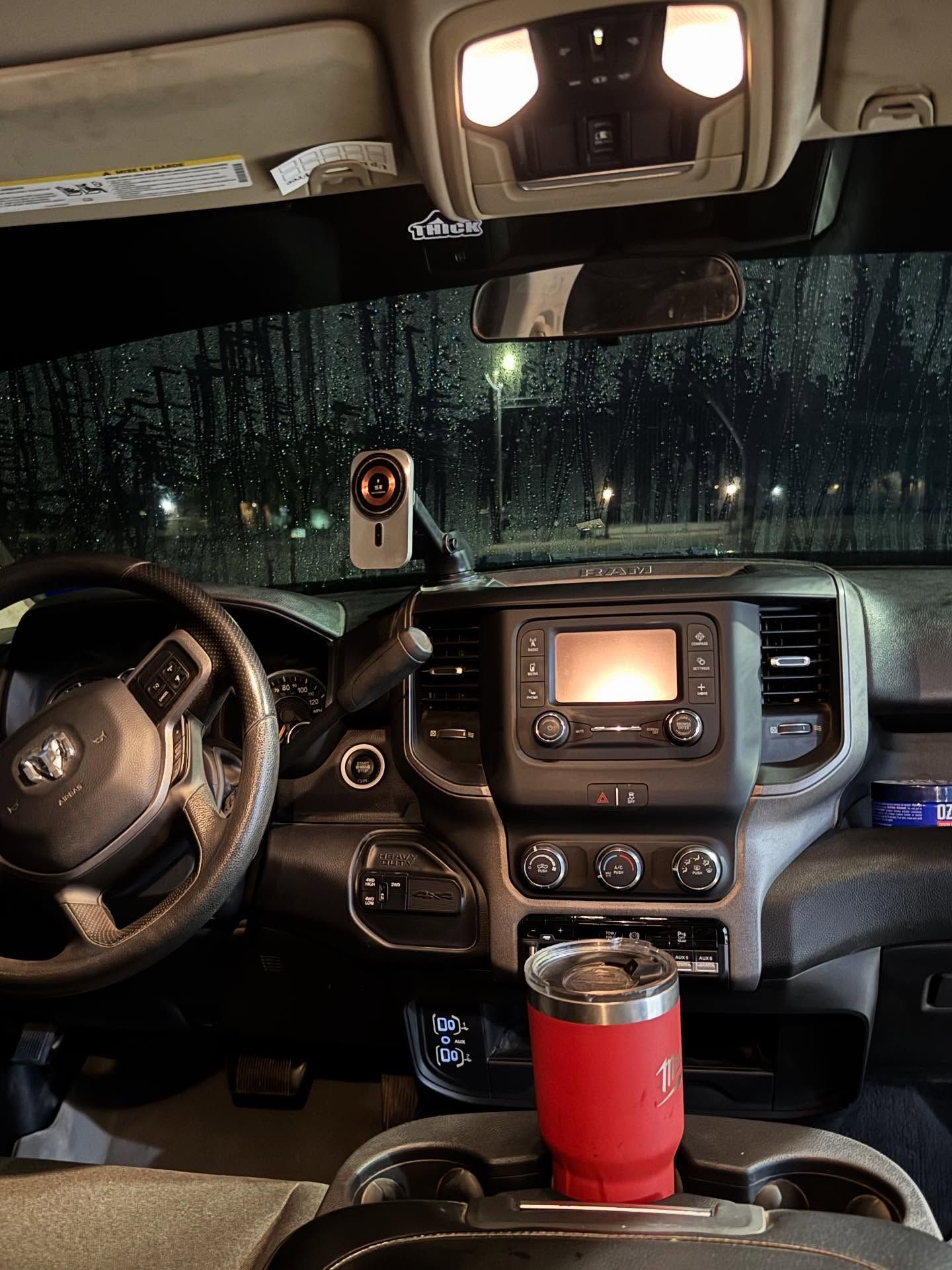 Truck cab interior with rain-streaked windshield, dashboard display, phone mount, and red tumbler in cup holder