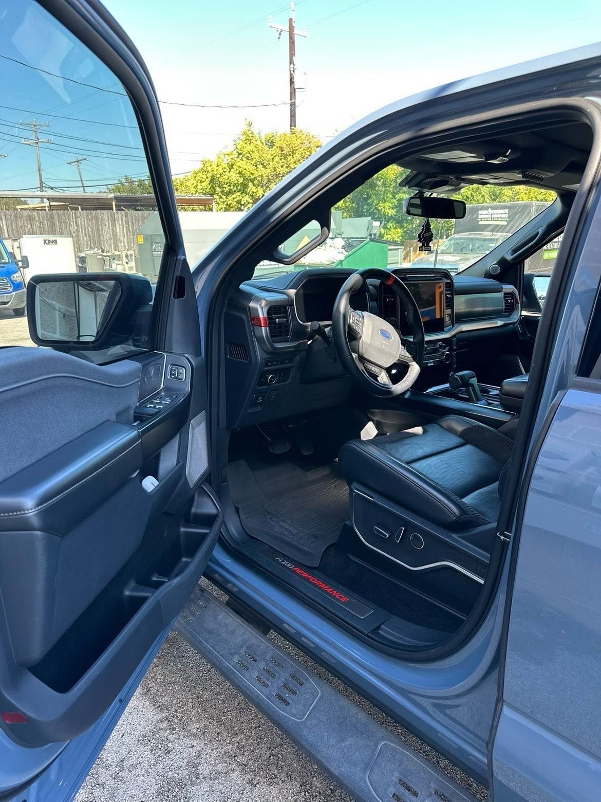 Open car door showing a steering aid device attached to the wheel and a black leather seat.