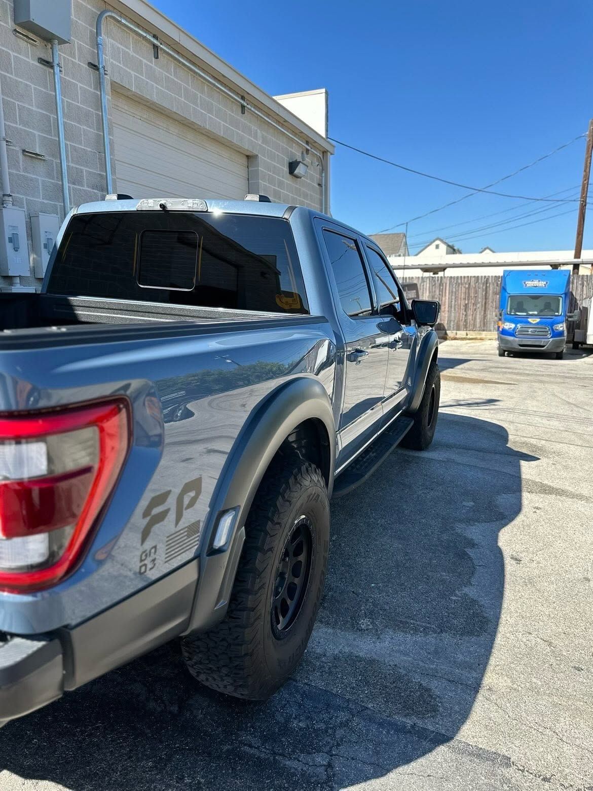 Gray pickup truck parked beside a building in a sunny lot, with a blue van in the background
