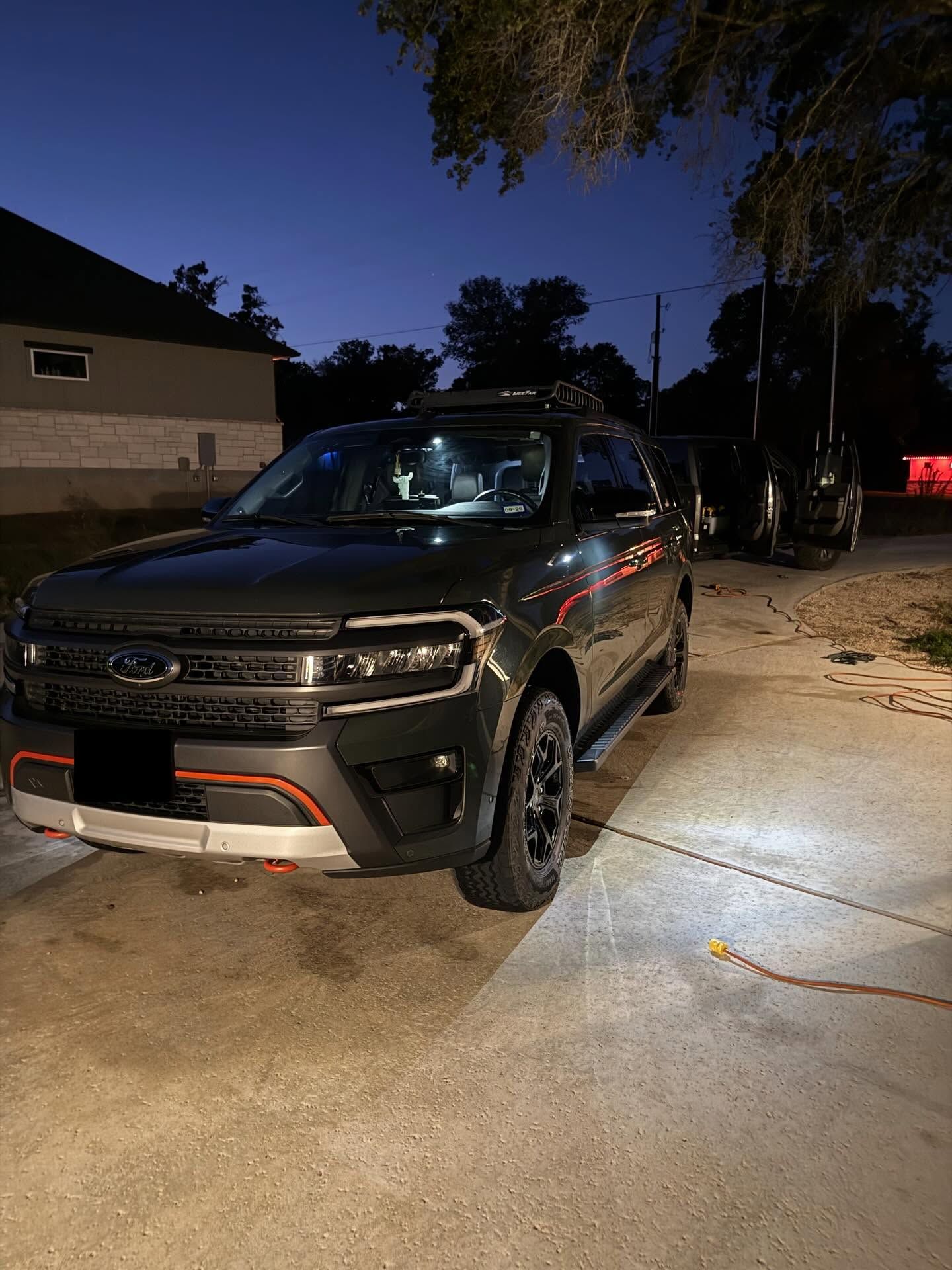 Dark SUV parked on a driveway at dusk, with headlights on and a house in the background.