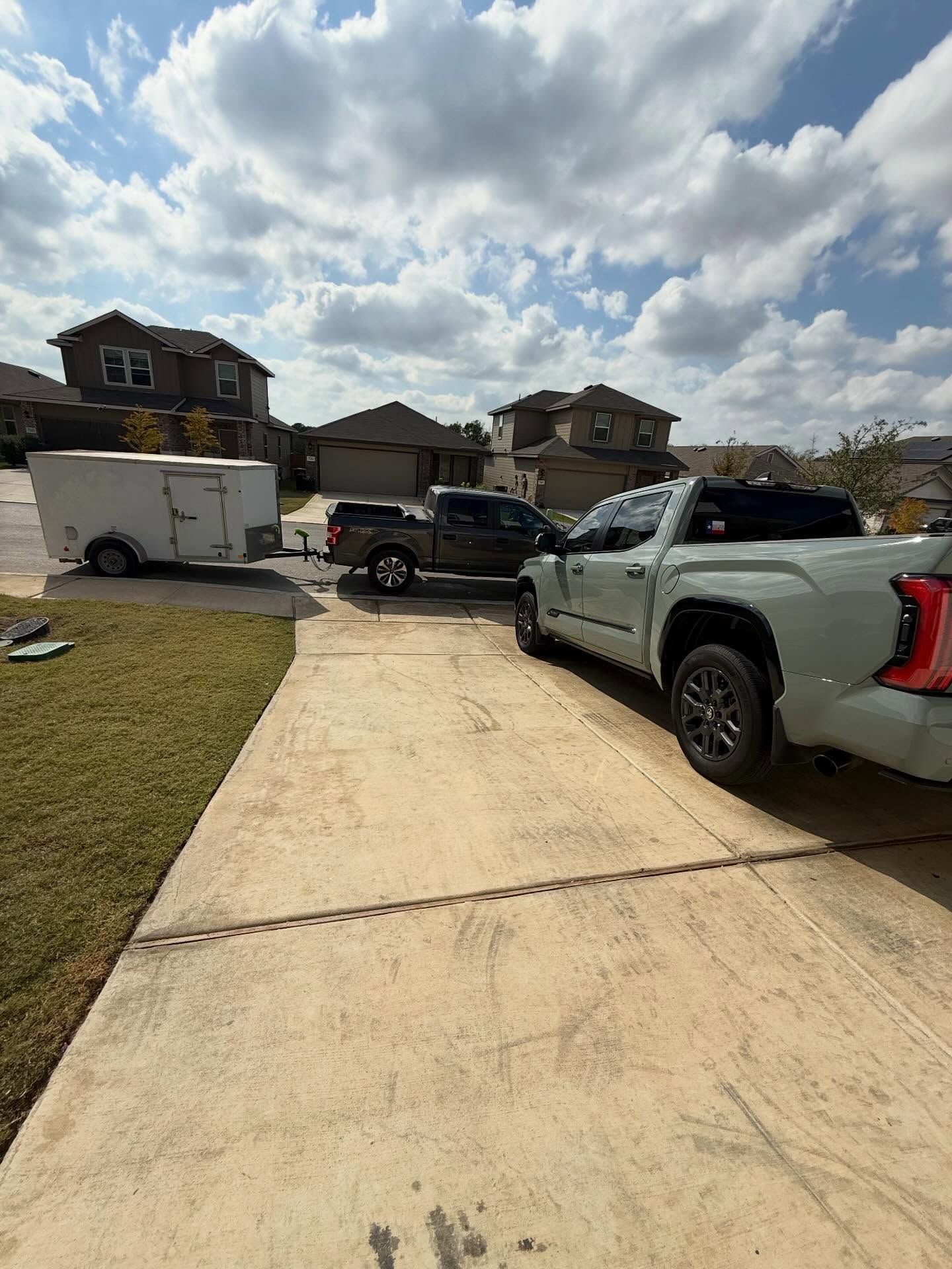 Driveway with several parked vehicles and a trailer in a suburban neighborhood under a cloudy sky.