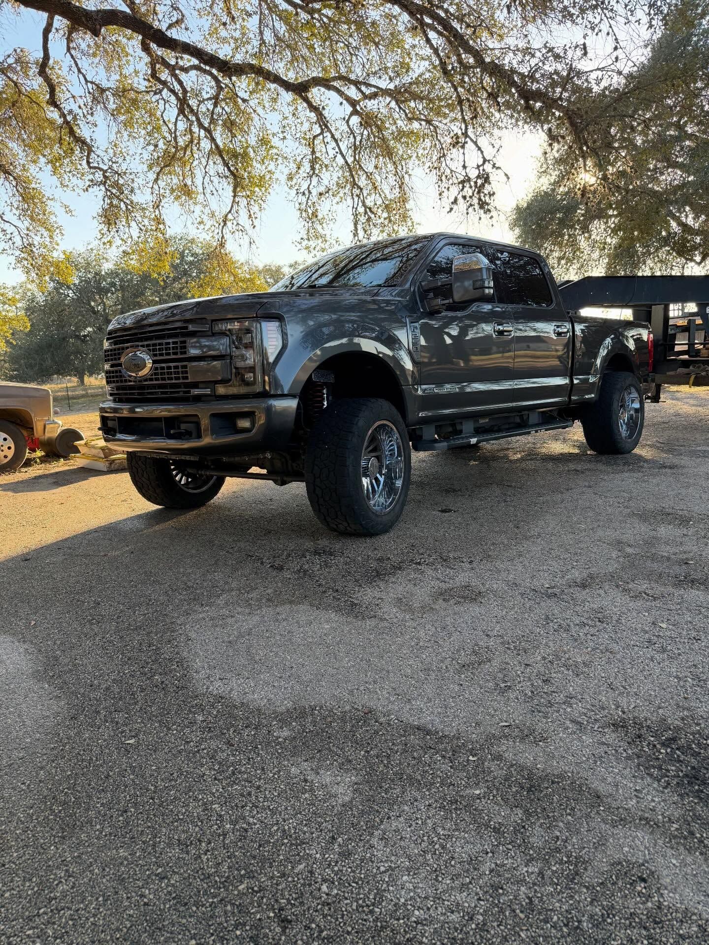 Dark gray lifted pickup truck parked on gravel under trees at sunset