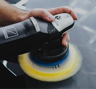 Hand using a black electric polisher with a spinning yellow-and-blue pad on a car surface
