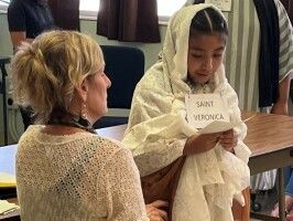Child in a white costume labeled “Saint Veronica,” standing beside a seated woman in a classroom or stage setting.