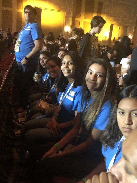 People in blue shirts seated at a crowded indoor event, some smiling at the camera.