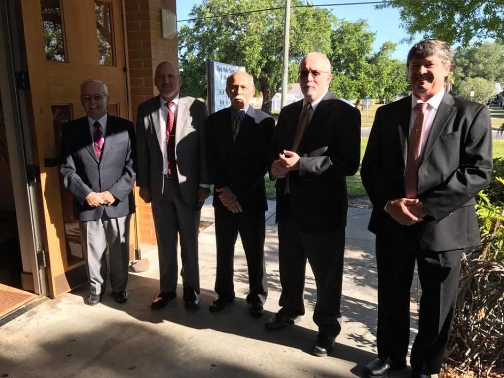 Five men in suits standing outdoors near a building entrance, in bright sunlight.