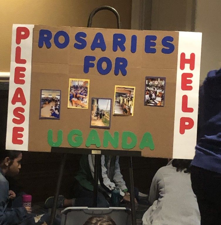 Sign reading “Rosaries for Uganda” with “Please” and “Help” on the sides, displayed at an event