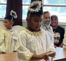 Children in white robes with gold trim stand indoors, one holding an object during a ceremony