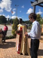 Outdoor ceremony with two people in robes and a man in a white shirt beside a flag and cross monument