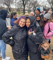 Two smiling people in black jackets pose outdoors with a group behind them on a cloudy day