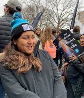 Woman in a blue beanie smiles at an outdoor protest, with other people and signs behind her.