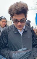 Man in dark jacket and glasses looking down at a phone, with another person behind him outdoors