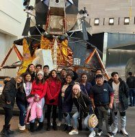 Group of people posing in front of a spacecraft exhibit in a museum, with metallic structure and a large dark display backdrop