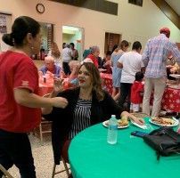 People mingling at an indoor event, with a woman seated at a green table holding a drink and food plates nearby.