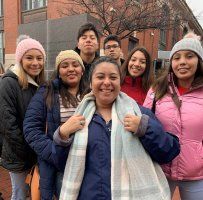 Five people smiling outdoors in winter coats, with a brick building in the background.