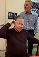 Two men indoors; one seated in a maroon sweater, the other standing behind him near a restroom sign.