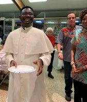 Person in white outfit holds a plate in a lobby while several people stand behind him.