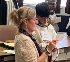 Two people in a classroom, one signing a paper while the other watches and holds a red pen.