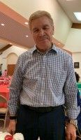 Man smiling in a plaid shirt and dark slacks, standing indoors near tables and festive decor.
