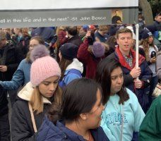 Crowded outdoor gathering with people in coats and hats, some holding signs, in a chilly setting