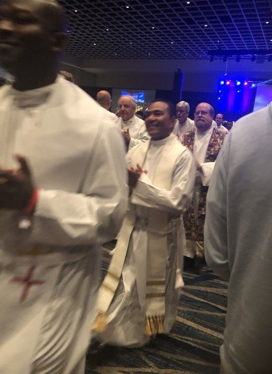 Clergy in white robes walking through a dimly lit hall with blue lights, some smiling and carrying programs.