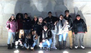 Group of people posing indoors against a white wall, bundled in coats and hats, smiling at the camera.