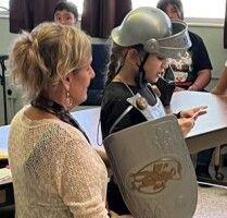Child in helmet and shield stands beside an adult in a classroom setting.