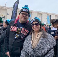 Two smiling people wearing winter hats and scarves outdoors at an event