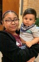 Woman holding a baby indoors, both looking at the camera, brick wall in the background